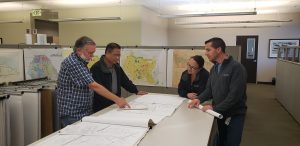 4 YLWD Team members reviews engineering plans on a large table. maps on cubicles surround them