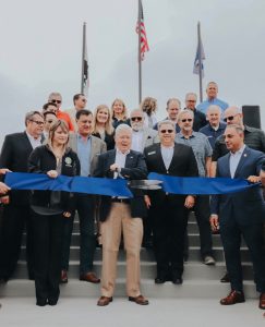 Director Brooke Jones cuts a large ribbon with large scissors. Dignitaries join him on a staircase with the US, CA, and YLWD flags in the background.