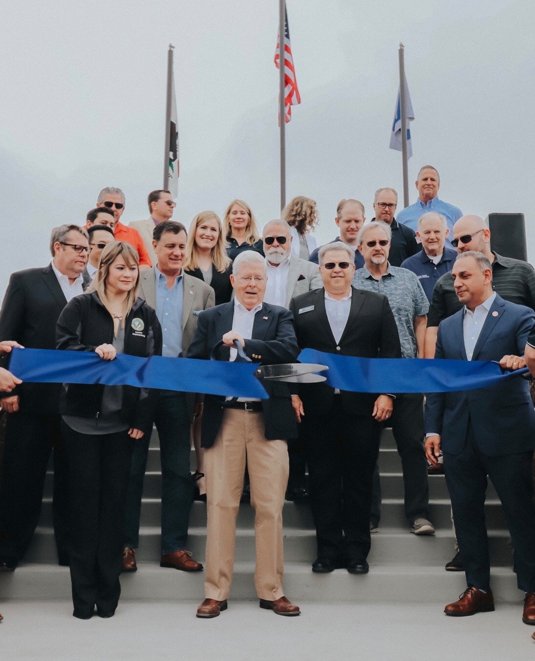 Director Brooke Jones cuts a large ribbon with large scissors. Dignitaries join him on a staircase with the US, CA, and YLWD flags in the background.