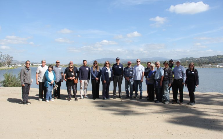 19 people take group photo standing on dirt in front of body of water