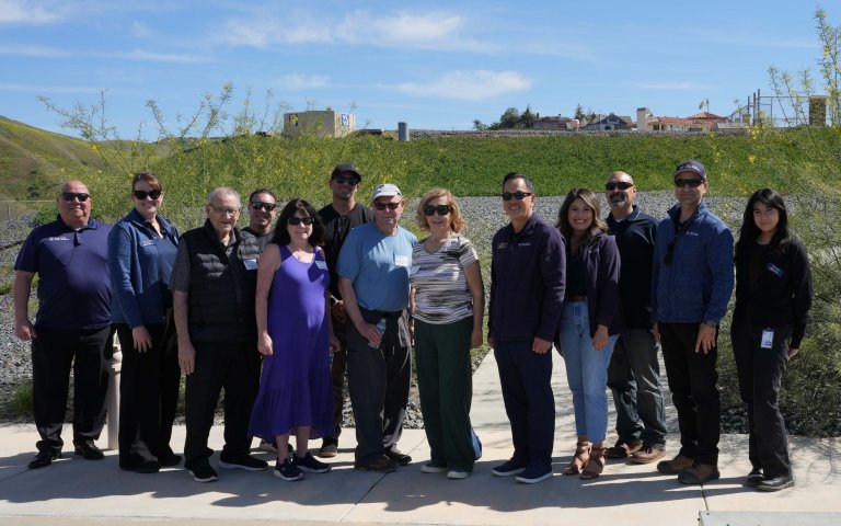 13 people pose for a group photo. The Marc Marcantonio Heli-Hydrant is in the background on a grassy hill behind group.
