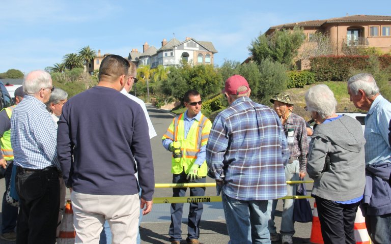 Maintenance worker holds flashlight pointing down towards opening in ground leading towards the sewer lift station. Tour group is standing around cones and caution tape in residential neighborhood in the middle of the street.