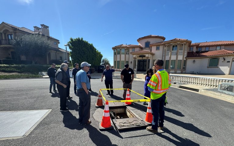 group is gathered at the end of a cul-de-sac in residential area, surrounding a square opening on the street with orange cones and caution tape
