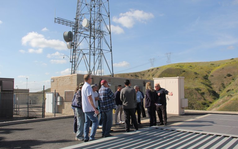 Group stands under satellite tower in front of brown building. The group is standing above light grey shafts near grassy hillside.