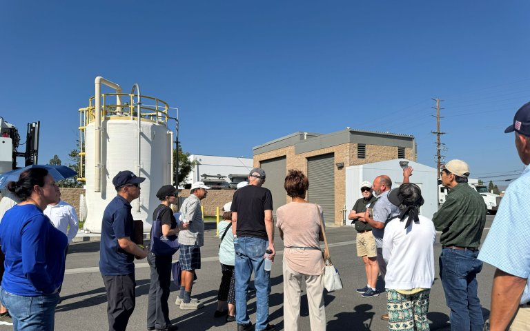 Group stands in front of large white cyclinder structure with yellow bars near brown building next in parking lot