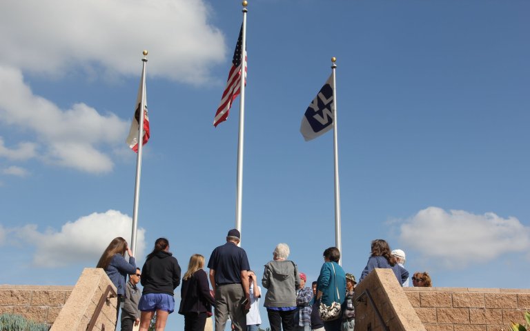 Group of people stand under three flag poles with California, US, and YLWD flags.