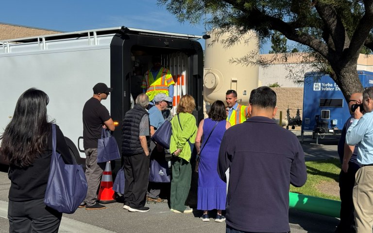 Tour group gathers by the back of an opened van near above ground sewer pipeline demonstration