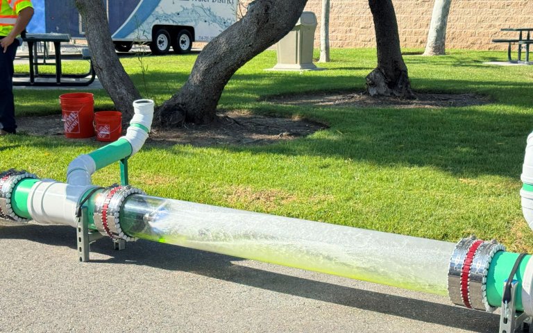 Clear horizontal pipe with green liquid and robotic camera inside spraying water, near grass and parking lot